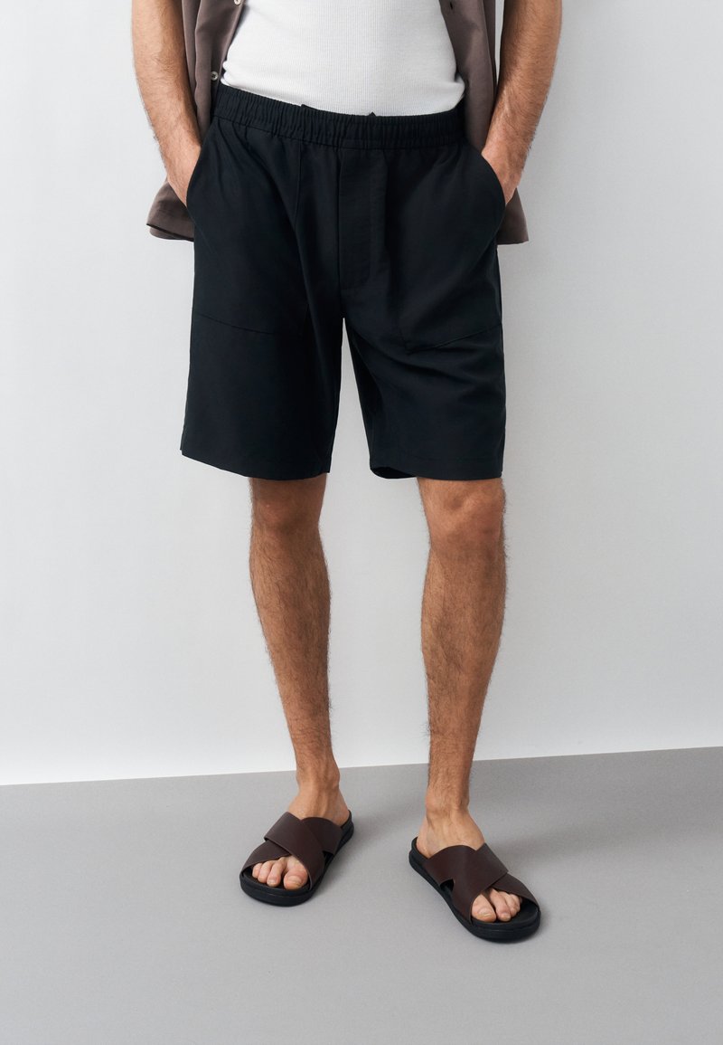 Homme portant un short à taille élastique noir, un débardeur blanc, des sandales marron à bouts ouverts, debout les mains dans les poches devant un fond uni.