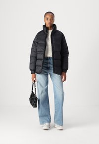 Casaco puffer preto com gola alta, tecido texturizado, fecho com zíper, combinado com jeans de perna larga azul claro e uma pequena bolsa preta.