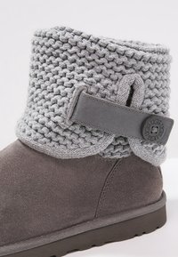 Botín de tobillo de ante gris con un grueso puño de punto y una correa decorativa que presenta un cierre de botón circular de marca.
