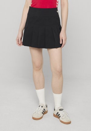 Schwarzer Faltenrock mit hohem Bund, aus glattem Stoff gefertigt. Kombiniert mit weißen Socken und klassischen mehrfarbig gestreiften Sneakers.