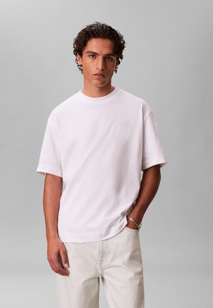 Jonge man met krullend haar die een effen wit T-shirt met korte mouwen en lichtbeige broek draagt, staand tegen een grijze achtergrond.