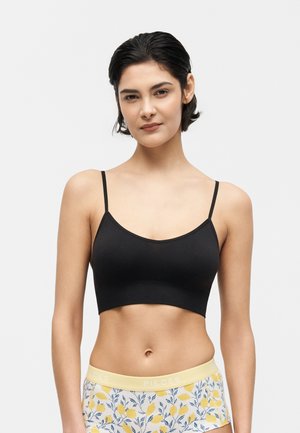 Žena s krátkymi tmavými vlasmi, oblečená v čiernom krátkom topu a žltých kraťasoch s kvetinovým vzorom, stojaca pred bielym pozadím.