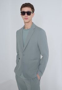 Giacca di completo grigio chiaro con tessuto testurizzato, un bottone e due tasche frontali, abbinata a una t-shirt grigio chiaro coordinata. Occhiali da sole neri indossati.