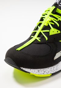 Scarpa sportiva nera con tomaia testurizzata, lacci verdi neon e dettagli. Presenta una suola bianca maculata con accenti neri.