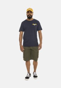 Camiseta de manga corta azul marino con logotipo y acentos amarillos, pantalones cortos cargo en verde oliva, y zapatillas negras con suelas blancas. Gorra amarilla en la cabeza.