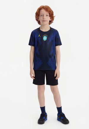 Κόκκινο αγόρι που στέκεται, φοράει σκούρο μπλε αθλητική φανέλα με το έμβλημα της Βραζιλίας, μαύρο σορτς, μπλε κάλτσες και μαύρα αθλητικά παπούτσια σε λευκό φόντο.