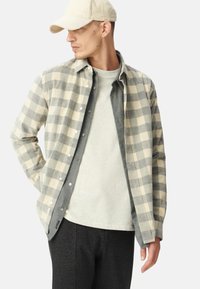 Chemise en flanelle à carreaux beige et gris, superposée sur un t-shirt gris, associée à un pantalon noir. Une casquette en velours côtelé beige vient compléter le look.