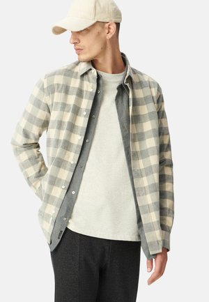 Geruite flanellen overhemd in beige en grijs, gelaagd over een grijs T-shirt, gecombineerd met donkere broek. Beige corduroy pet maakt de look compleet.