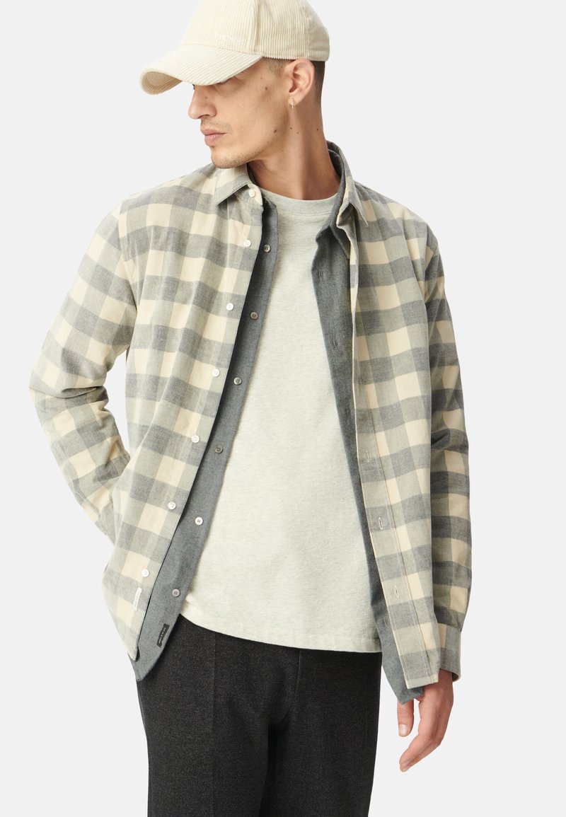 Chemise en flanelle à carreaux beige et gris, superposée sur un t-shirt gris, associée à un pantalon noir. Une casquette en velours côtelé beige vient compléter le look.