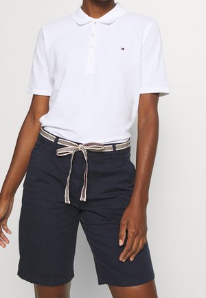 Person wearing white short-sleeve polo shirt, navy blue shorts with a patterned fabric belt tied in a bow at the waist.