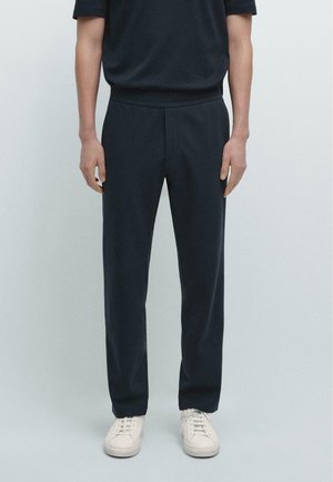 Uomo con pantaloni neri a gamba dritta e sneakers bianche, in piedi davanti a uno sfondo chiaro e uniforme.