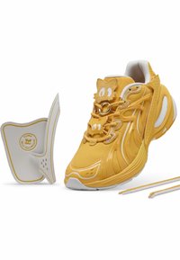 Chaussure de sport jaune avec des superpositions en mesh et synthétiques, dotée d'accents blancs, d'éléments décoratifs en forme d'animaux sur la languette, et de lacets supplémentaires.