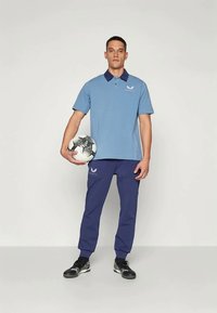 Μπλε πόλο μπλούζα με ναυτικό γιακά, κεντημένο λογότυπο, συνδυασμένη με ναυτικές φόρμες και μαύρα αθλητικά παπούτσια, κρατώντας μία ασπρόμαυρη μπάλα ποδοσφαίρου.