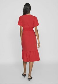 Červené šaty do délky kolen s krátkými rukávy, s nabíraným pasem a vrstveným volánkovým lemem. Hladká textura látky. Nosené s černými sandály.