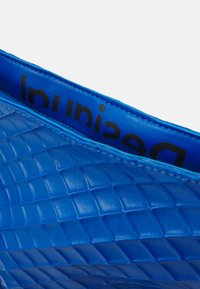 Blaues strukturiertes Lederetui mit einem gesteppten Muster. Das Innere bietet ein glattes blaues Futter und schwarzen Logotext. Mit Reißverschlussverschluss.