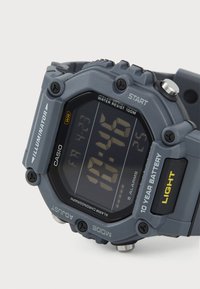 Montre numérique avec un boîtier en plastique gris, un cadran noir, un grand écran LCD, trois boutons, étanche jusqu'à 100 mètres et cinq réglages d'alarme.