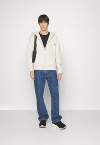 Haut gris clair à fermeture éclair avec poches avant, porté sur un t-shirt noir, associé à un jean bleu et des baskets noires. Tissu lisse, design décontracté.