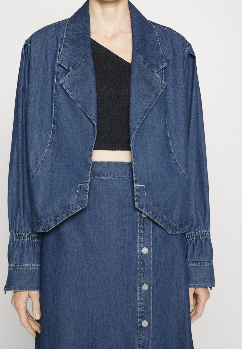 Jeansjacka med en kortad design, breda slag och elastiska ärmslut, kombinerad med en svart smockad topp och en matchande jeanskjol.