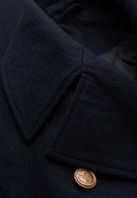 Marineblauer Wollmantel mit strukturierten Oberflächen, großem Kragen und bronzefarbenem Knopfdetail mit kunstvollem Design.