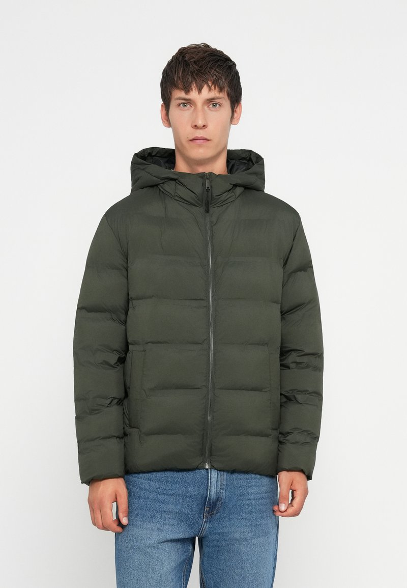 Giovane uomo che indossa una giacca imbottita verde scuro con cappuccio e zip frontale, abbinata a jeans blu, in piedi contro uno sfondo bianco uniforme.