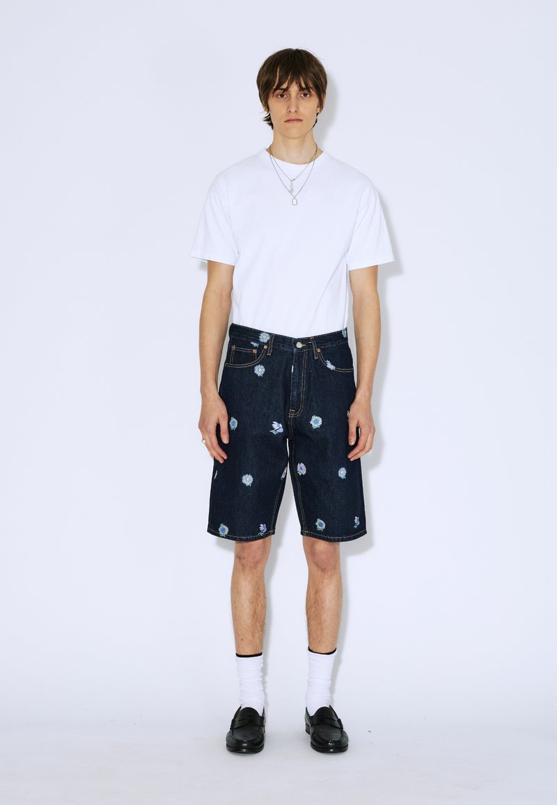 Pantalones cortos de mezclilla con bordado floral, color azul oscuro, corte regular, longitud a mitad del muslo, combinados con una camiseta blanca lisa y zapatos negros.
