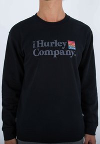 Černá mikina s nápisem "THE Hurley Company" v tučném šedém písmu, s barevným obdélníkovým logem vlevo. Jednoduchý design, bez vzorů.