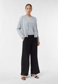Lichtgrijze cardigan met knopen, gecombineerd met een grijs vest en zwarte wijd-uitlopende broek. Zwarte open-teen hakken maken de outfit compleet.