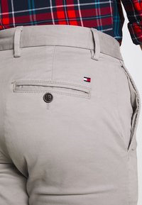 Pantalones de gris claro con un bolsillo trasero con cierre de botón y un pequeño logo bordado en rojo, blanco y azul. Textura de tela suave.