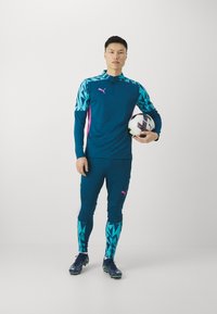 Blaues sportliches Langarmshirt mit türkisfarbenen geometrischen Mustern und pinken Akzenten, kombiniert mit passenden Hosen und schwarzen Fußballschuhen. Hält einen Fußball.