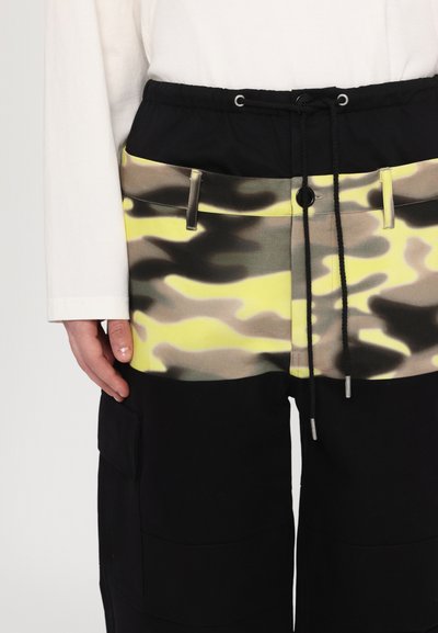 Pantaloni cargo negri cu un buzunar cu imprimeu camuflaj galben și gri, talie elastică cu șnur de strângere și două buzunare frontale. Textura materialului este netedă.