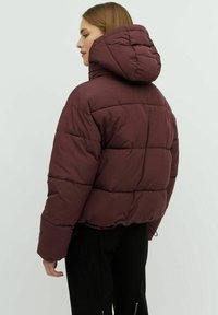 Giubbotto imbottito bordeaux con cappuccio, caratterizzato da un design a trapunta e una vestibilità oversized. Il tessuto appare liscio con una leggera lucentezza.
