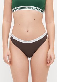 Rudas medvilninis thong tipo maudymosi kostiumas su švelnia elastinga juosta, ant kurios yra baltas "Lacoste" logotipas. Sukurtas su žemu juosmeniu ir sklandžia tekstūra.