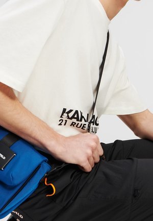 Hvid T-shirt med sort tekst, blå skuldertaske og sorte bukser. Tasken har en lynlås og orange detaljer. Fokus på teksturer og farver.