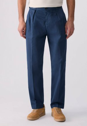 Homme portant un pantalon droit bleu marine et des chaussures en daim marron clair à lacets, debout devant un fond uni.