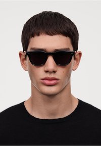 Ochelari de soare negri cu lentile întunecate și un cadru elegant. Accente argintii notabile la temple, purtați de o persoană într-un tricou negru cu guler rotund.