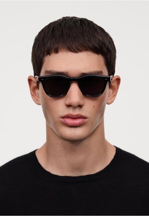 Ochelari de soare negri cu lentile întunecate și un cadru elegant. Accente argintii notabile la temple, purtați de o persoană într-un tricou negru cu guler rotund.
