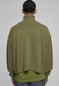 Pull oversize vert olive avec un col montant, des manches en maille torsadée texturée et un ourlet côtelé, porté par-dessus une chemise verte.