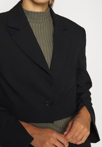 Veste noire coupée, col cranté, bouton noir unique et poches latérales. Portée sur un col roulé côtelé vert olive.