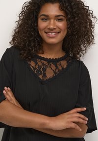 Haut noir avec une insert en dentelle à motif floral au niveau du col, manches courtes et tissu texturé avec des rayures verticales.