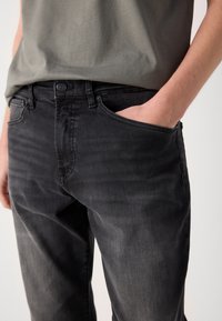 Personne portant un jean noir délavé avec une main dans la poche avant et une chemise grise rentrée de façon lâche à la taille.