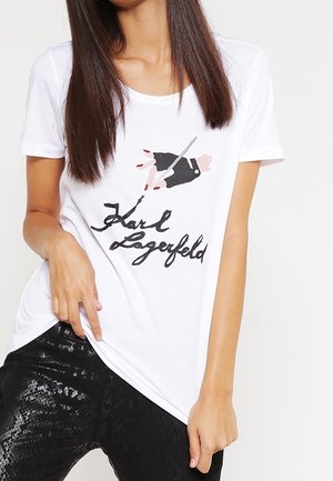 Frau trägt weißes T-Shirt mit Grafik einer handschuhbewehrten Hand, die einen Stift hält und „Karl Lagerfeld“ in schwarzer Schrift unterschreibt, kombiniert mit schwarzen strukturierten Hosen.