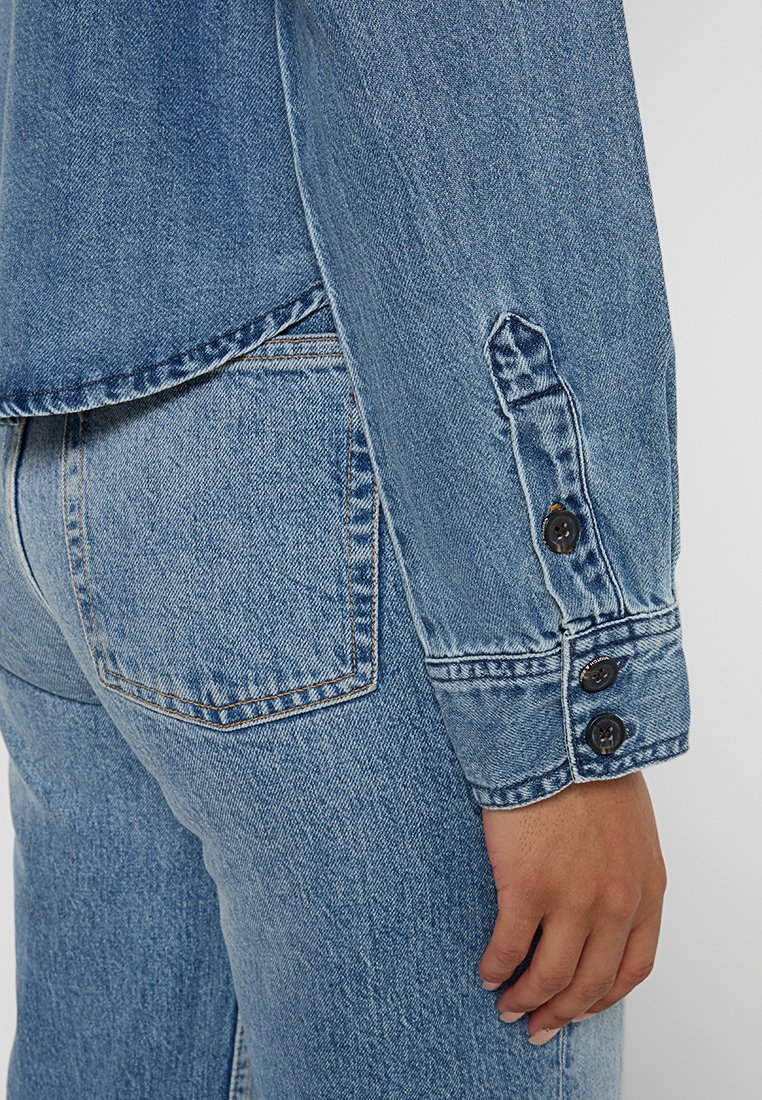 Leren spijkerjack in lichtblauw. Heeft een kraag, lange mouwen met knopen bij de manchetten, en een borstzak. Zichtbare stiksels op de randen.