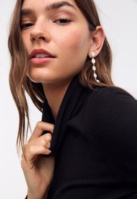 Zwarte trui met een hoge, geribbelde kraag. Model draagt hangende oorbellen met ovale, parelachtige kralen. Glad, schoon huid en subtiele make-up.