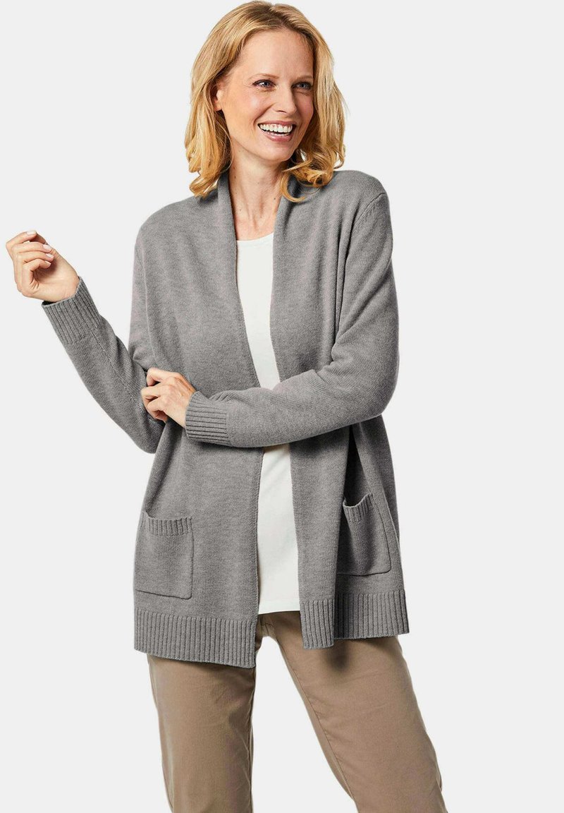 Grau Cardigan aus weichem Material, mit Schalkragen, gerippten Bündchen und zwei aufgesetzten Taschen vorne. Getragen über einem weißen Oberteil.