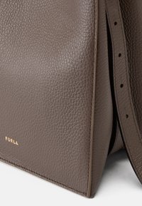 Bolso de mano de cuero marrón con una textura en relieve, que cuenta con un logo dorado, una base plana y una correa de hombro desmontable con agujeros ajustables.
