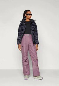 Fekete pufis dzsekit, fekete felsőt, laza lila cargo nadrágot, bézs tornacipőt és fekete napszemüveget viselő nő áll egy sima világos háttér előtt.