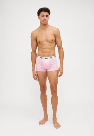 Boxer shorts rosa con una cintura logo nera, tessuto morbido, design aderente e copertura minima, indossati da un modello maschile in piedi.