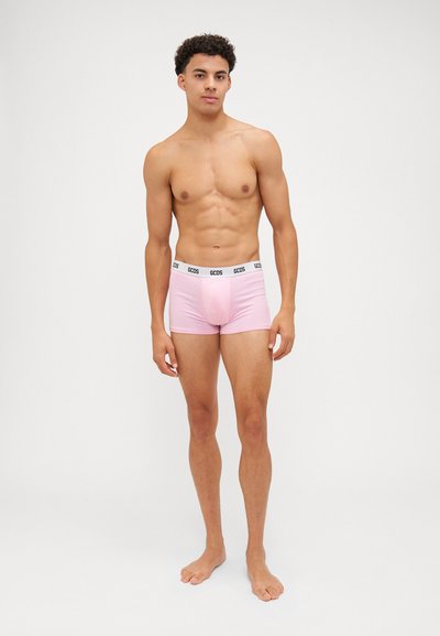 Boxer shorts rosa con una cintura logo nera, tessuto morbido, design aderente e copertura minima, indossati da un modello maschile in piedi.