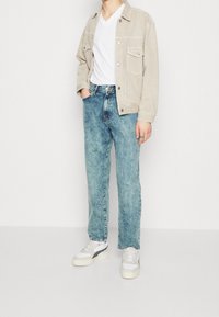 Chaqueta de ante beige con dos bolsillos en el pecho, camiseta blanca de cuello en V, jeans de mezclilla azul claro y zapatillas blancas con acentos grises.