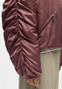 Bomberjakke med rynkede ermer i burgunder satin, med glidelåslomme på siden og ribbestrikkede mansjetter. Teksturert finish som forbedrer det overordnede designet.